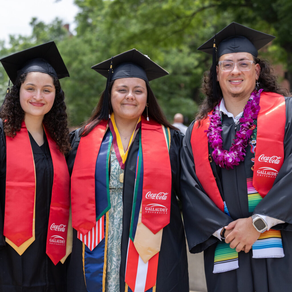 Three Gallaudet graduates smile for the camera. They wear graduation regalia, and each have distinctively red stoles that have the Coca-Cola and Gallaudet logos.
