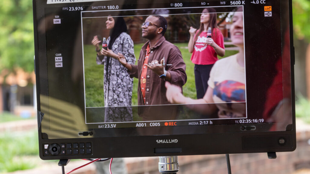 A photo of a camera monitor showing four Gallaudet students signing on a grassy area. The students are looking offscreen to the left, smiling and signing "today". Two are holding Coca-Cola bottles.