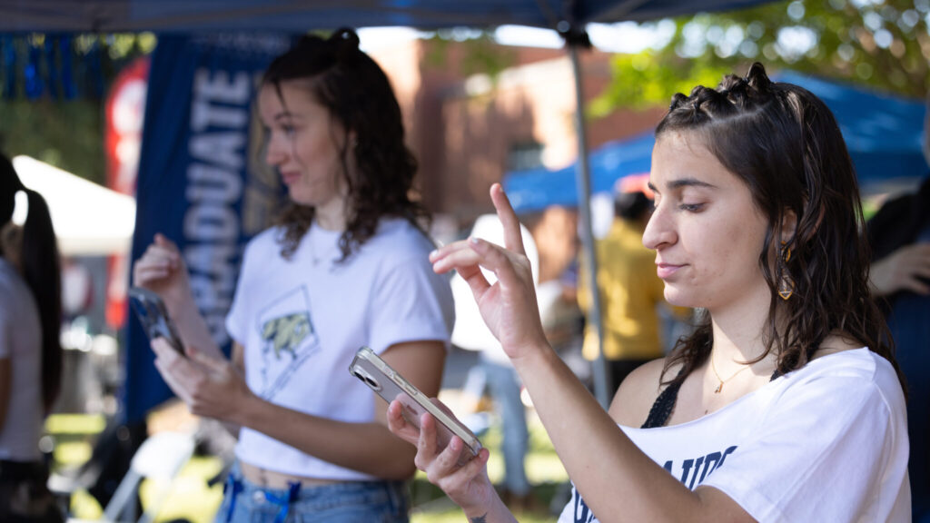 Two students in a booth at Gallaudet Homecoming. The student in the foreground, on the right, is looking down at their phone while signing. The student on the left, out of focus in the background, is holding their phone and looking to the left at a person offscreen.