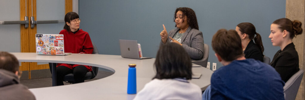 A photo of students presenting during a meeting. Six people sit around a curved table. Two students, on the left and in the center, are most visible, with laptops open in front of them. The student on the left is wearing a stark red top, looking to the right at the student in the middle. The middle student signs to their laptop. Two more students sit to the right, their faces partially obscured. Two more people are in the foreground, watching the middle student with their backs to the camera.