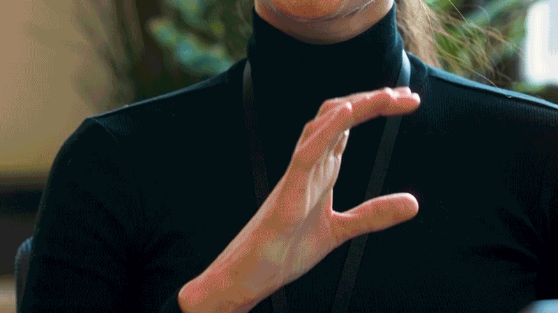 An animated gif of a hand fingerspelling C-O-K-E along the axis of the ribbon on Coca-Cola branding. The beginning shows a person's torso at a computer reaching for a bottle of Coca-Cola. As their hand makes the sign for "C", the image becomes an animation, with a white hand traveling along the ribbon, fingerspelling, before reverting back to the real person's hand holding the "E". Captions at the bottom say, "C-O-K-E spelling out 'Coke'".