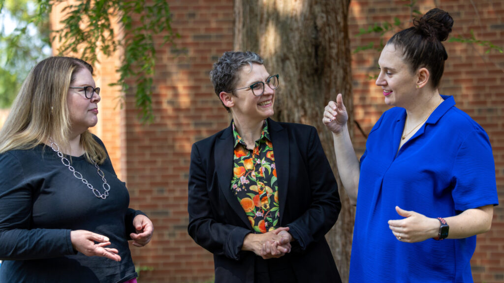 A photo of three people standing outside, looking at each other and smiling. On the left is a person with light brown hair past their shoulders, a black shirt, and necklace. In the middle is a person with short brown hair, a black blazer over a patterned shirt, and glasses. On the right is a person with dark brown hair in a bun and a royal blue polo. In the background are a brick wall and trees.