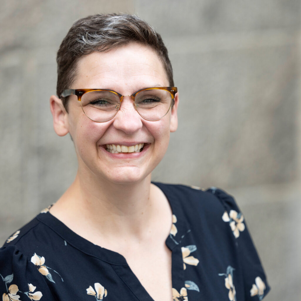 A headshot photo of person smiling brightly. They have short brown hair and glasses. They wear a patterned shirt.