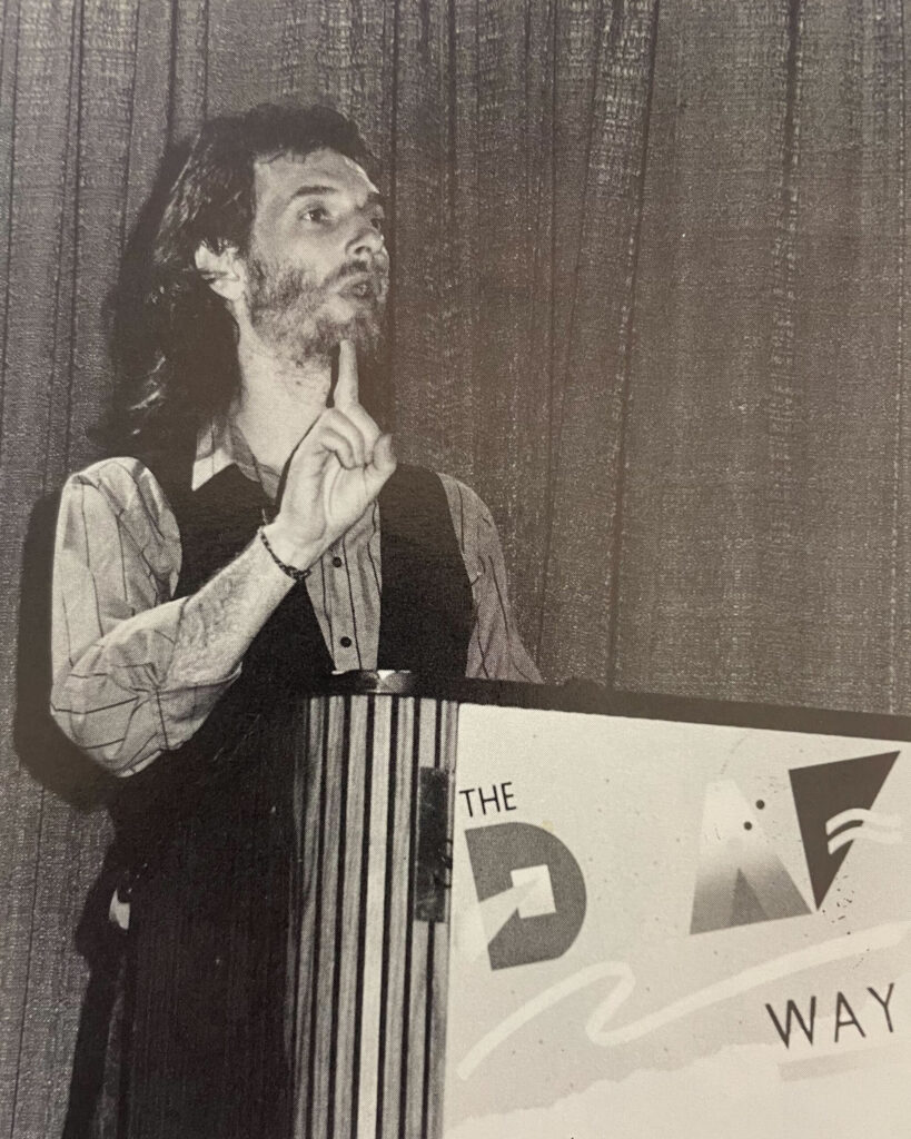 A black-and-white photo of Paddy Ladd giving a lecture at Deaf Way I. Ladd is standing at a podium, signing to the right at an audience offscreen. He has dark hair to his shoulders and a light beard, with a dark vest over a striped shirt. The podium, to the bottom right, has a Deaf Way logo.