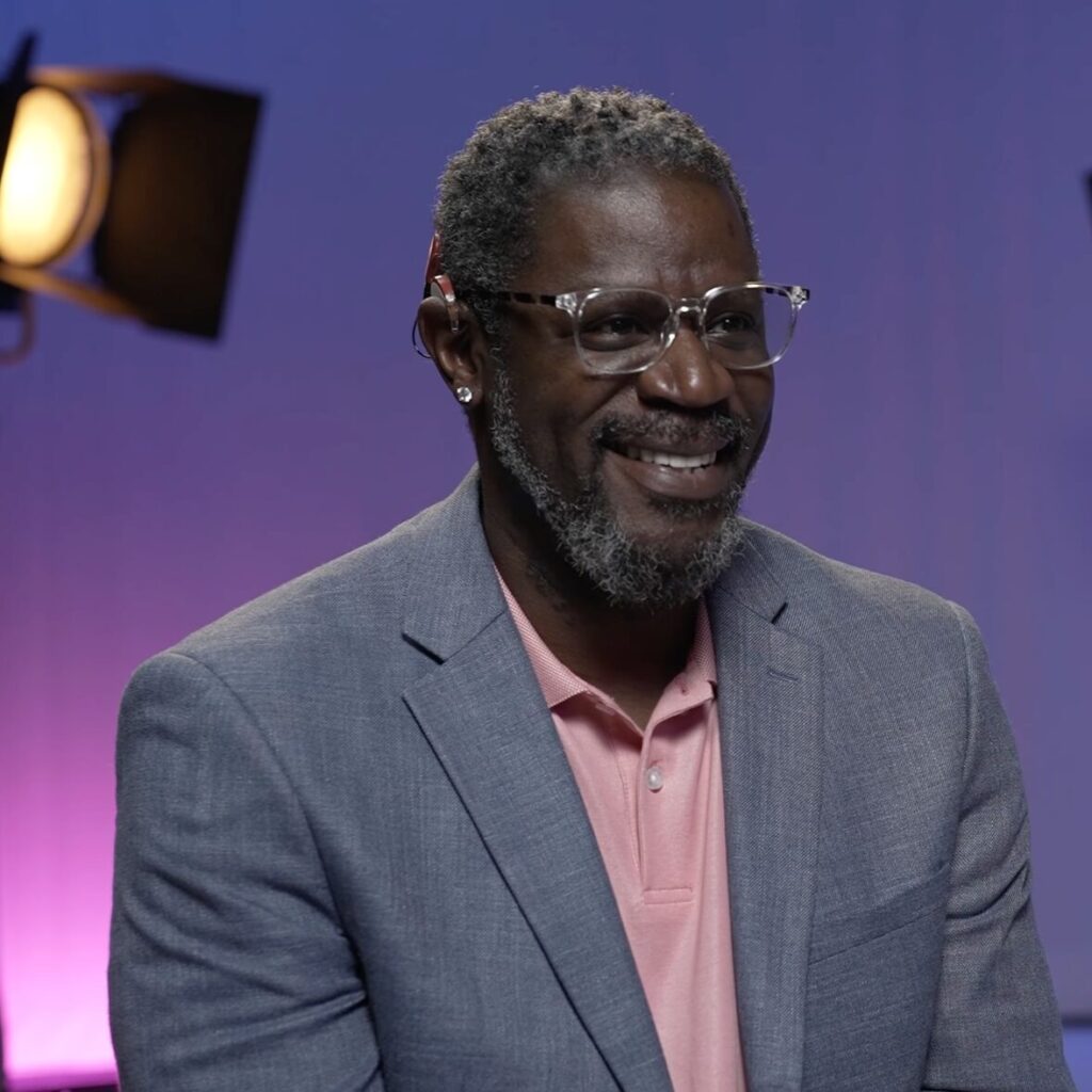 A photo of Warren "Wawa" Snipe during an interview. He is smiling at a subject offscreen to the right. He is wearing a glasses, a cochlear implant, and a blue-gray suit over a pink polo. The background is light Gallaudet blue, with a  filming light visible in the top left corner.