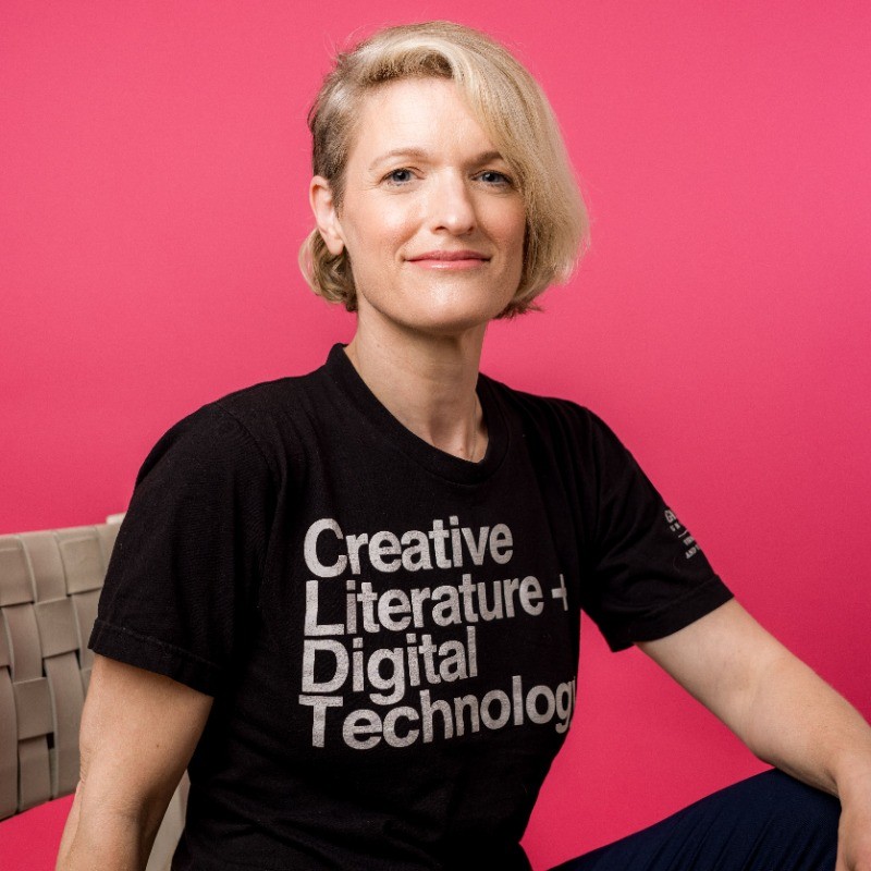 A photo of Melissa Malzkuhn sitting in front of a pink background. She is striking a confident pose, with her left arm on a crossed leg, and shoulders diagonal to the camera. She has blonde past her ears. styled to cover the left side of her head. She is wearing a black tee that says, "Creative Literature + Digital Technolog".