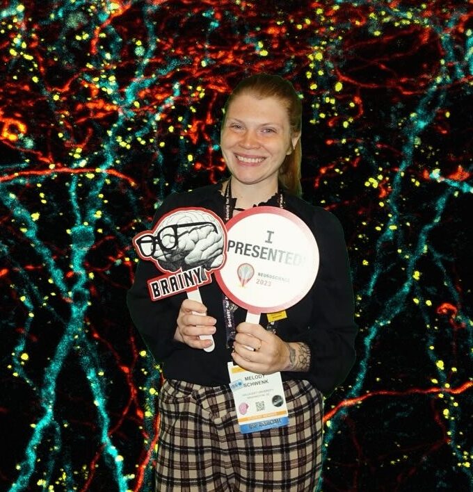 Woman stands in front of a black backdrop decorated with lots of different colored lights. She is holding two paper fans. One has an image of a brain wearing glasses and the other reads, "I Presented." She is wearing a conference badge and smiling.