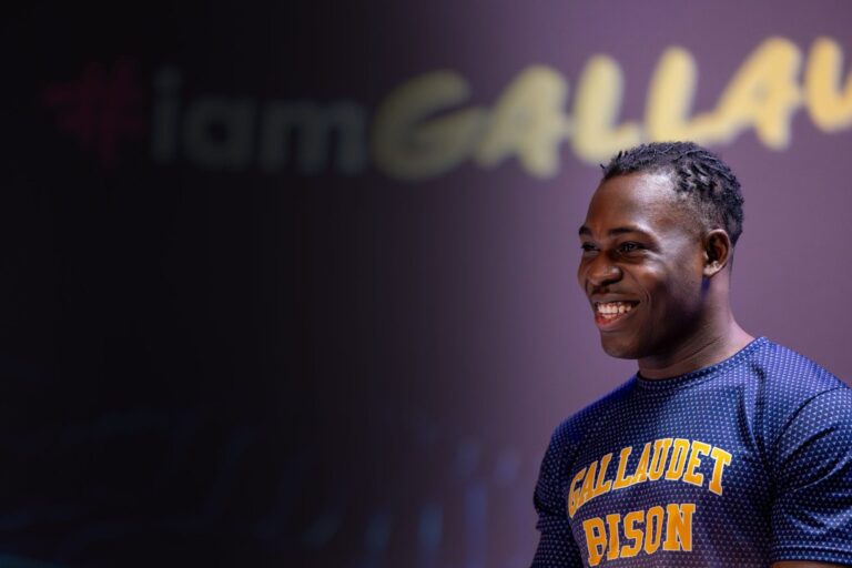 A smiling male athlete wearing a blue and yellow "Gallaudet Bison" sports shirt, with a blurred background featuring the words "iamGallaudet." The atmosphere is positive and energetic.