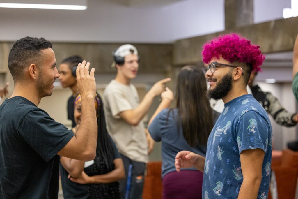 A group of people engaged in conversation in a casual setting. Two individuals in the foreground are smiling and interacting, with one person having pink hair and wearing glasses. Others are seen in the background, some pointing and laughing, creating a lively atmosphere.