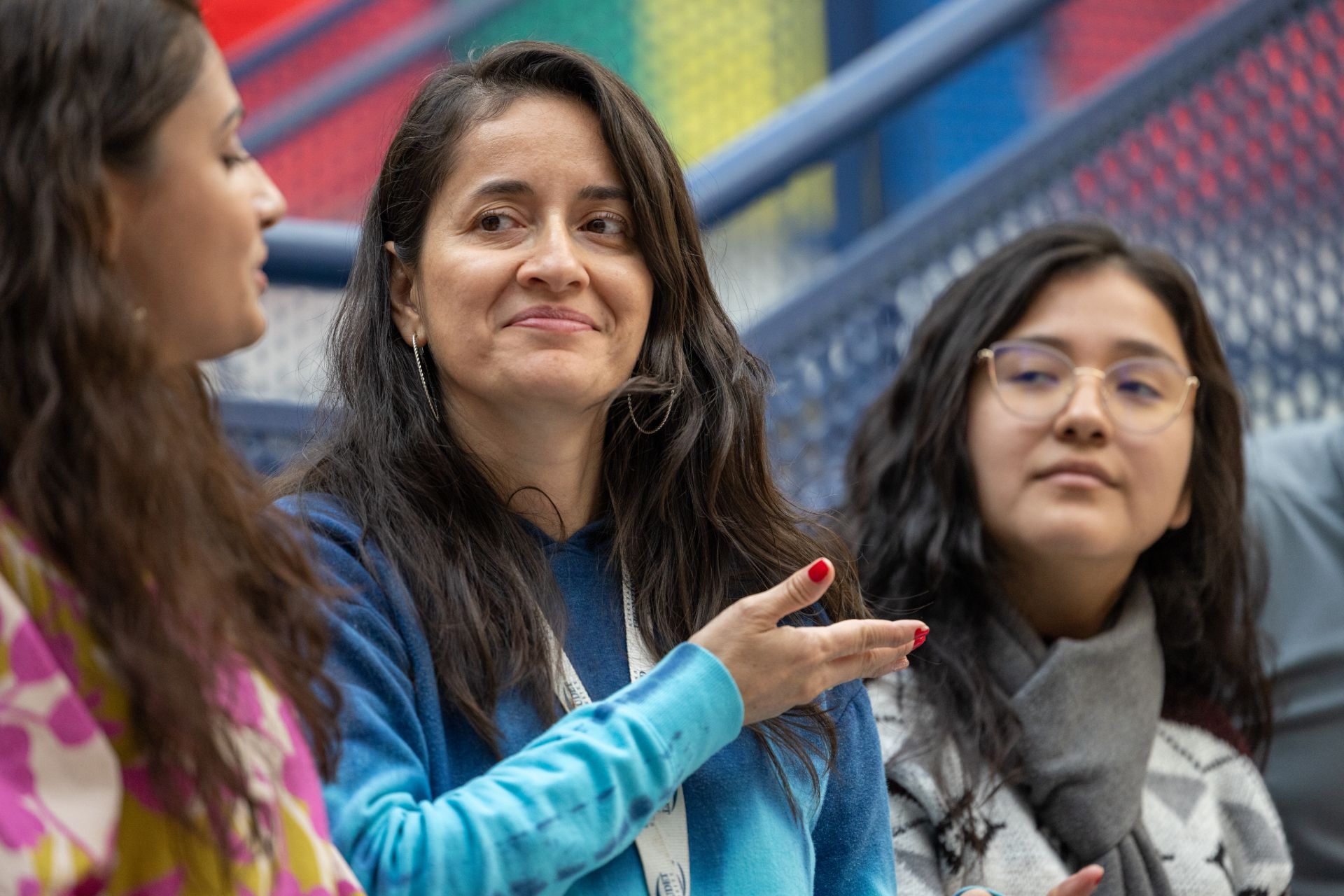 Three women are engaged in conversation. One woman, wearing a blue top, is smiling and gesturing as she speaks. The others are listening, with one in a colorful patterned shirt and the other in a gray scarf. The background features colorful geometric patterns.