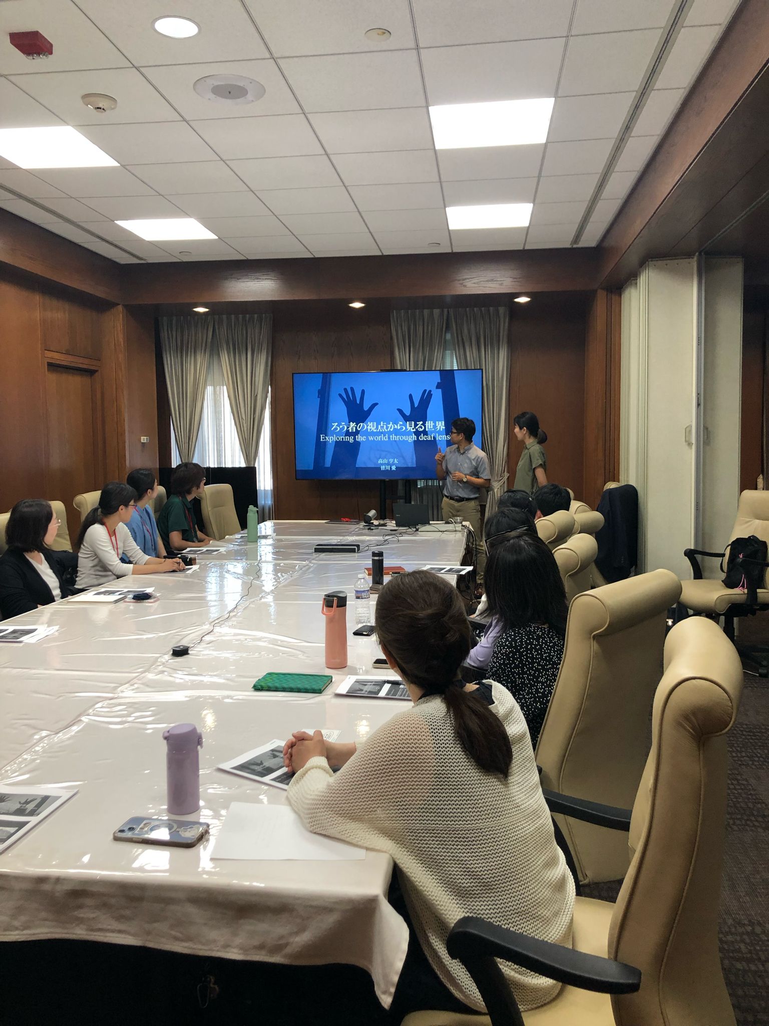 A group of people are seated around a rectangular conference table. At the end of the table, a man and a woman are standing in front of a screen showing the image of hands, Japanese text, and the words, "Exploring the world through deaf lens."