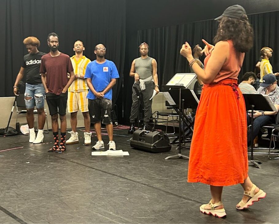 A photo of a theatre rehearsal in a room with black walls and floor. Five actors in the back look to the right foreground at a director wearing a red dress and black hat. The back right wall of the room is a mirror. Music stands and two other seated people are visible.