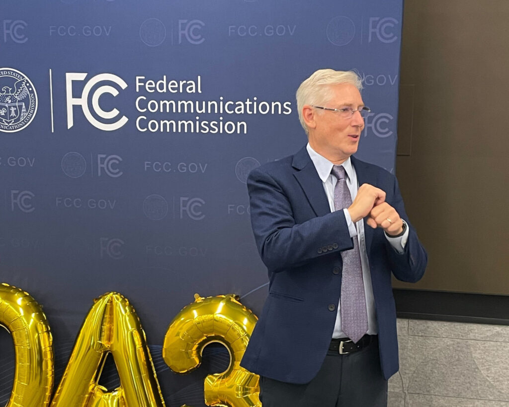 Greg Hlibok signs to the right at a person offscreen. Greg has short white hair and glasses, and wears a blue suit with a tie. Behind him is a blue backdrop with a logo and name: "Federal Communications Commission". The FCC logo on the backdrop is also on the award. On the ground against the backdrop are golden balloons in the shape of letters and numbers.