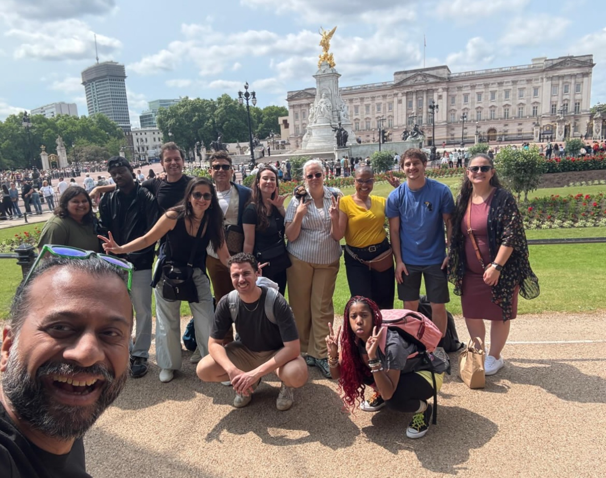 A man takes a selfie photo that features a group of people posing behind him. They are outside on a beautiful sunny day in a grassy area with a grand white building behind them.