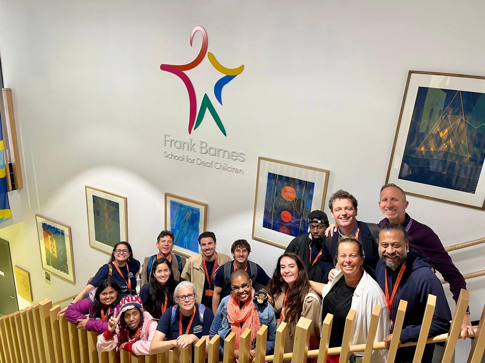 Group of people pose while standing on a staircase. Behind them are several colorful posters and the words "Frank Barnes School for Deaf Children."
