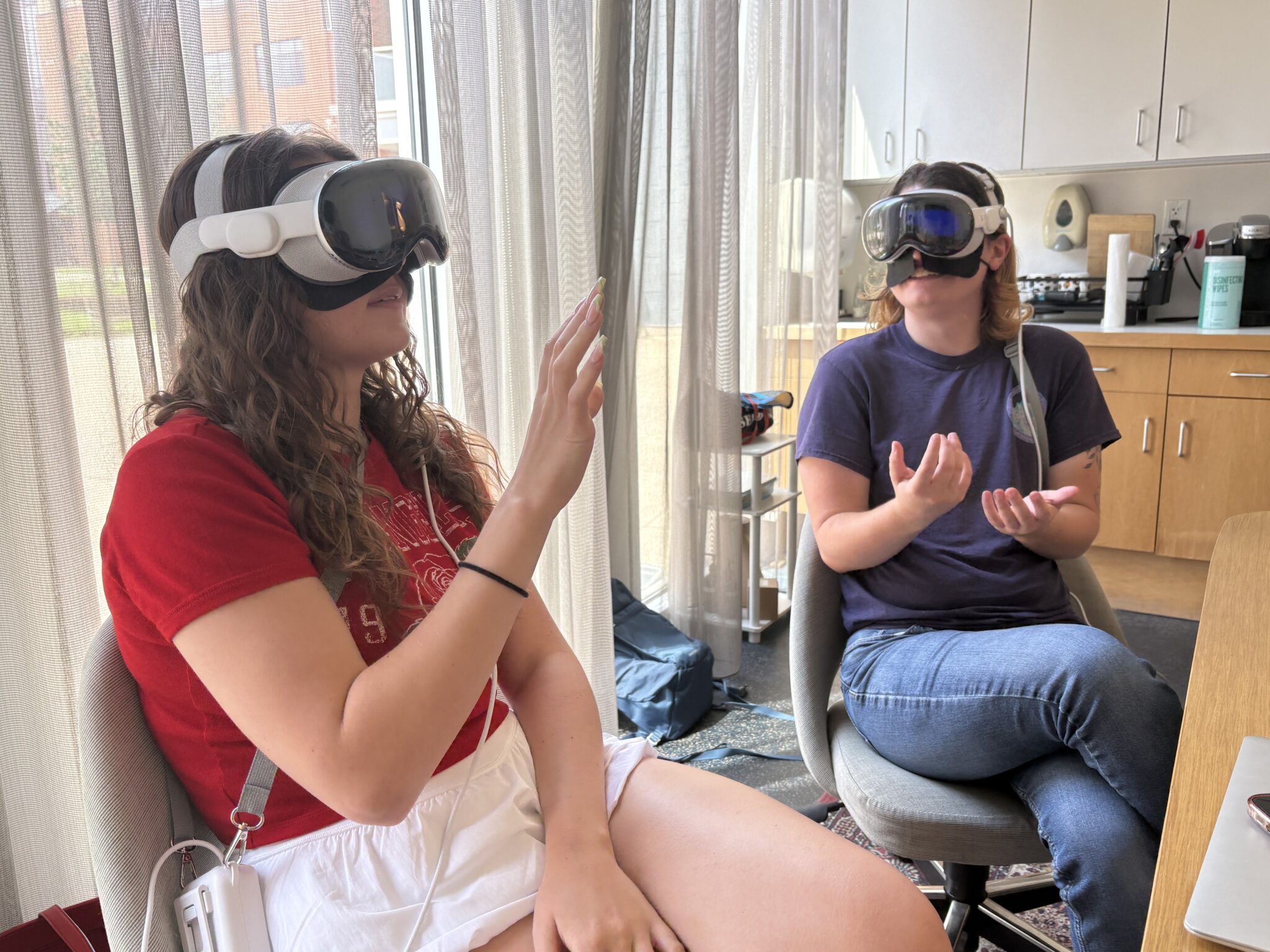 Two women are seated next to each other while wearing VR headsets. They are both making movements with their hands.