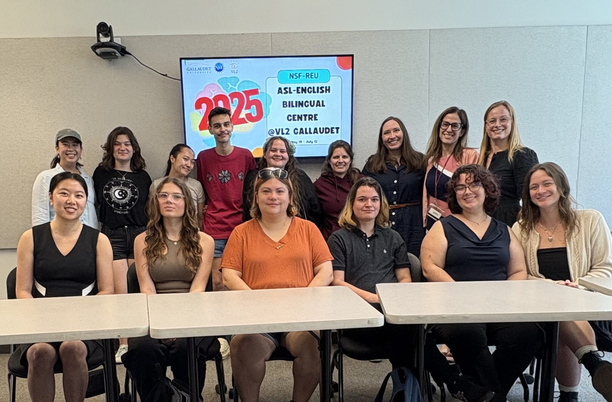 About a dozen people pose for a group photo. One row is seated at two white tables while another row stands behind them. A screen behind the group reads, "ASL-English Bilingual CENTRE @VL2 Gallaudet"