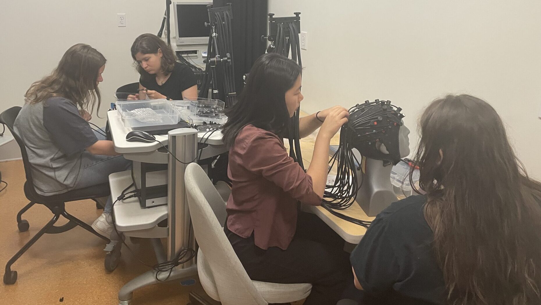 Four women are working at a set of tables. They are working with various kinds of testing equipment. The most prominent is a black cap that is on a mannequin's head. One woman is manipulating the wires coming off it.