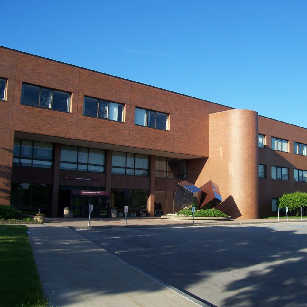 A photo of the National Technical Institute for the Deaf (NTID). A brick building in front of a concrete parking lot and sidewalk. On the right side of the building's face is a projected section shaped like a book sticking out of a bookcase. To the bottom right of the projected section is a sculpture that looks like a large, shiny, black Rubik's cube split vertically down the seams.
