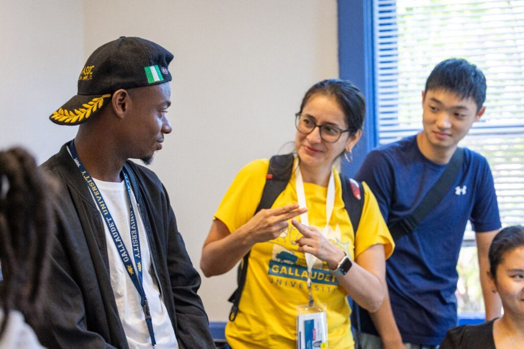 A group of three young adults engaged in conversation in a bright room. One person is wearing a yellow t-shirt and glasses, animatedly talking to another individual who has a black cap and a black jacket. The third person, wearing a dark blue shirt, is listening attentively. The setting appears casual and friendly.