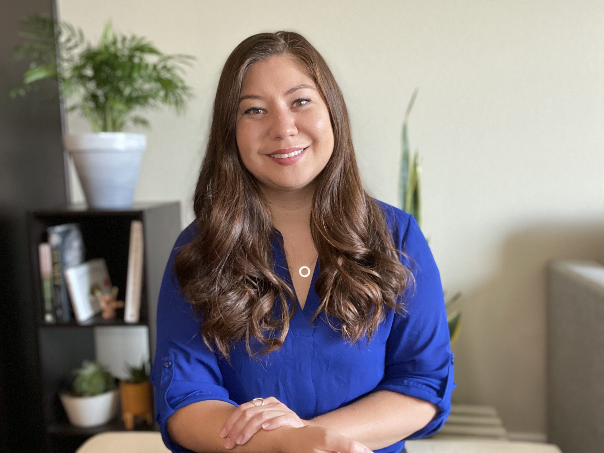 Woman in blue blouse with long hair poses at a desk with a plant behind her