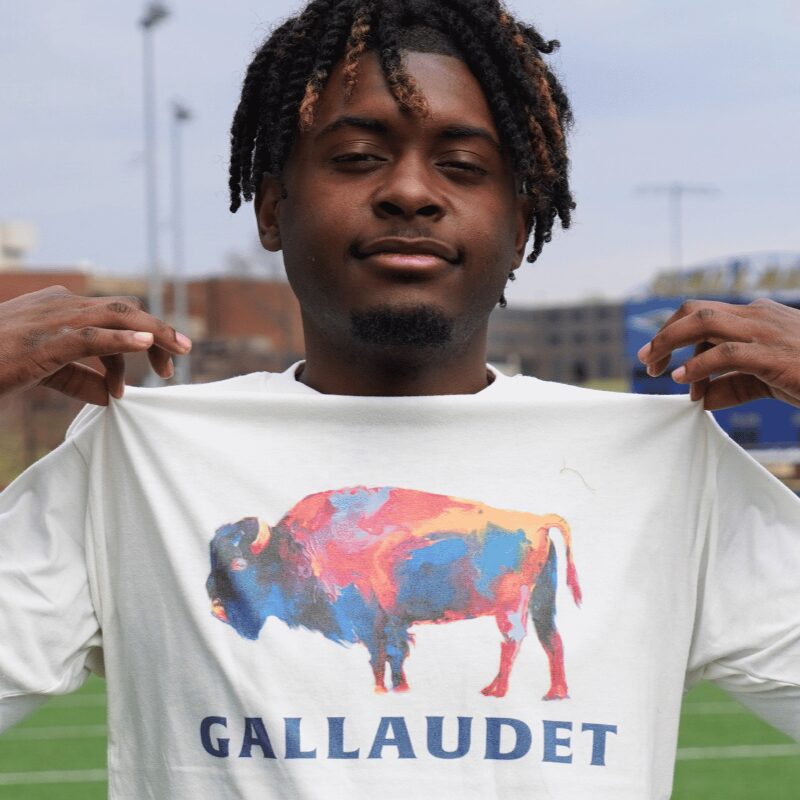 A photo of a person standing and pulling out the chest of their white t-shirt, which has a colorful Bison over the word "Gallaudet". They are standing on a football field, blurry in the background.