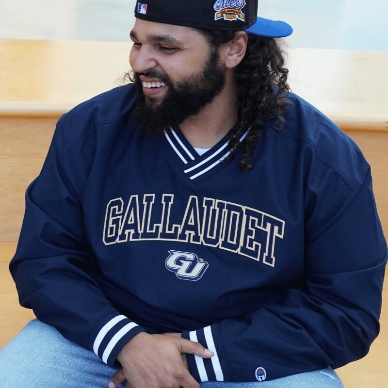 A photo of a seated smiling person looking to the left. They are wearing a dark blue baseball pullover jacket and a backwards cap.