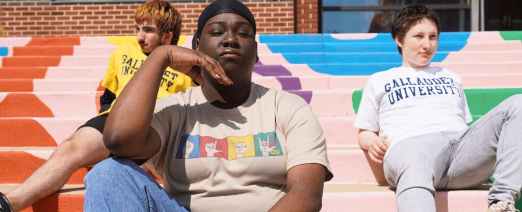 A photo of three people sitting on a wide set of outdoor stairs, wearing Gallaudet merchandise. Prominent in the foreground is a person with their head resting casually on their right hand. They wear a shirt with the ASL handshapes for "I", "L", "U", and "I love you". In the background are two people with "Gallaudet University" shirts.