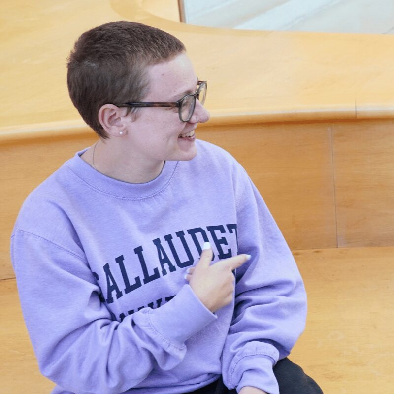 A photo of a seated smiling person looking to the right. They are wearing a light blue sweatshirt with obscured text that can be assumed to read "Gallaudet University".