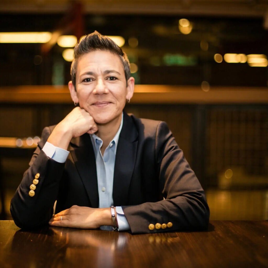A photo of Dr. Maribel Gárate-Estes sitting with her head on her right fist, smiling lightly at the camera. Her dark hair, flecked with gray, is short and styled into a peak in the front. She is wearing a dark blazer over a light blue button-up shirt, and earrings. The blazer has four golden buttons on the back of each wrist. The background is a blurred business room.