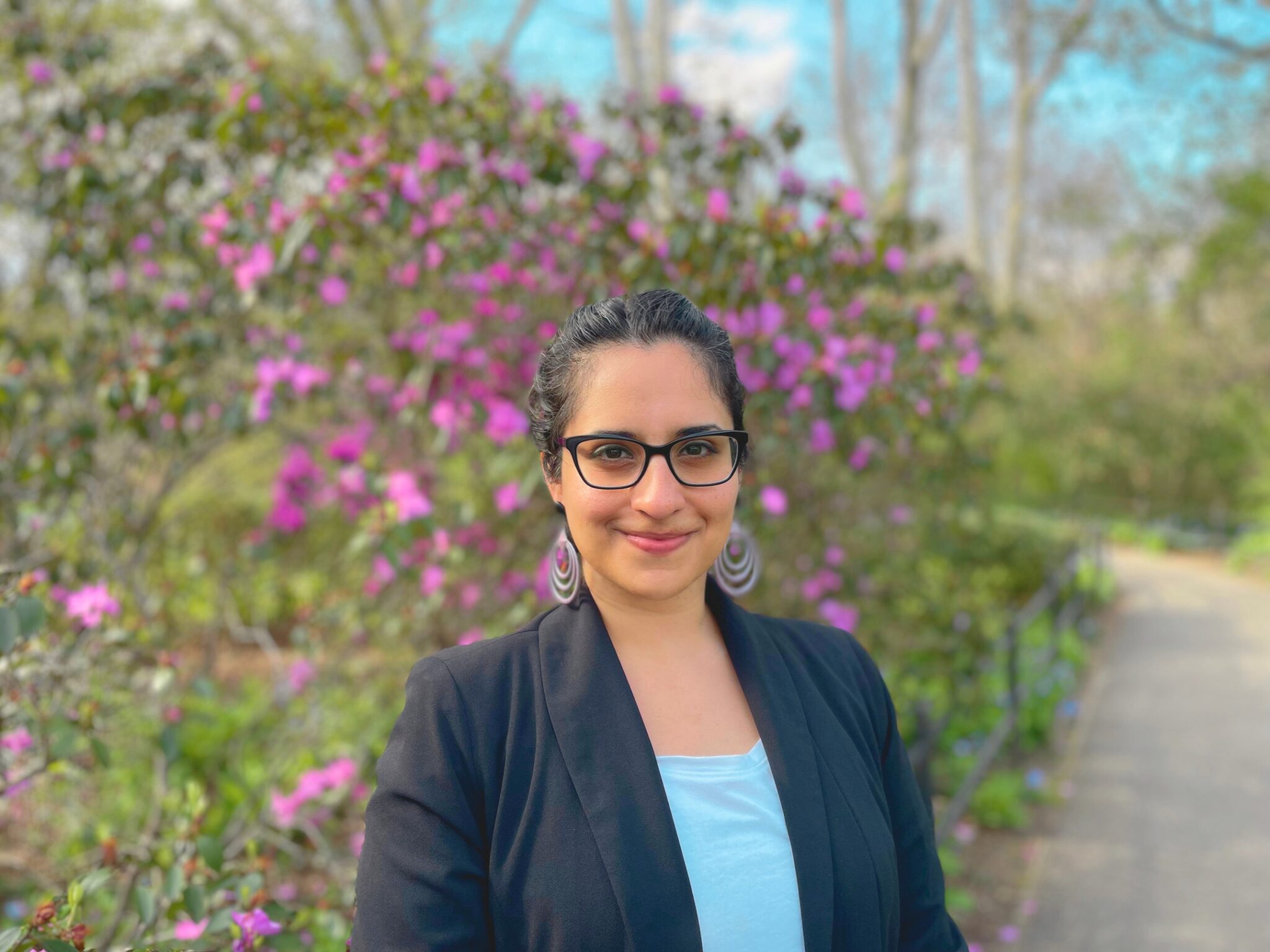 Woman in glasses wears a light blue shirt with a jacket over it and stands in front of a large bush covered in purple flowers