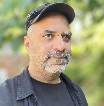 Man with facial hair wears a black baseball cap, a black T-shirt, and a black collared shirt