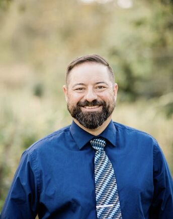 Man with facial hair wears a blue collared shirt with a striped tie and poses outside with trees and plants behind him