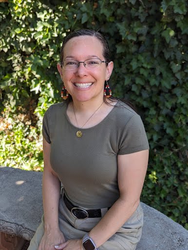Woman in glasses wears a dark green shirt and sits on an outdoor bench in front of plants