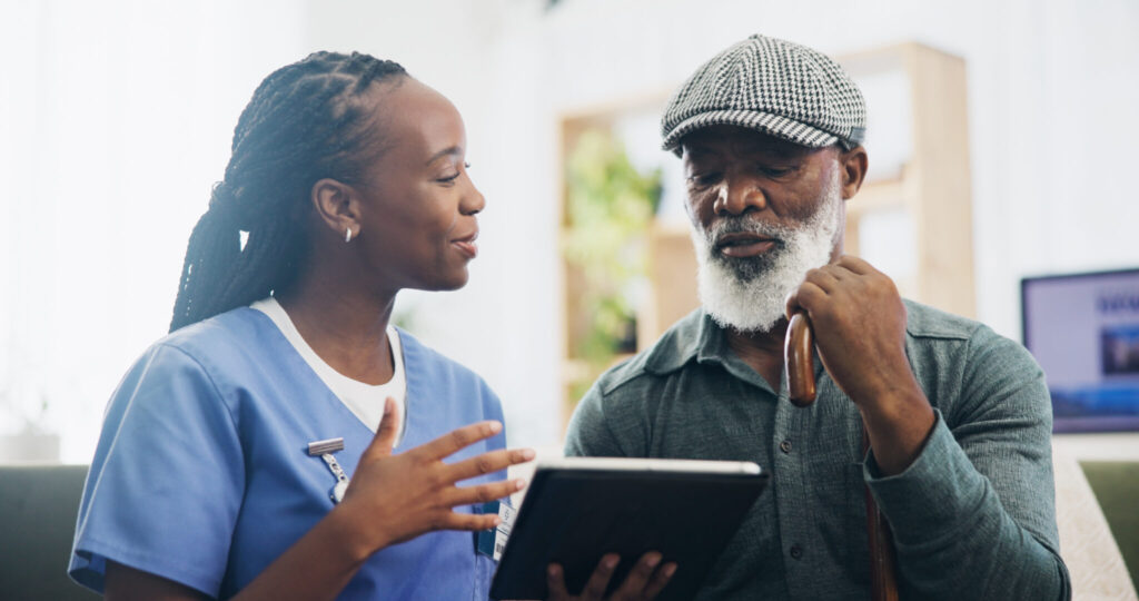 A healthcare professional in scrubs is engaged in a conversation with an elderly man who is holding a cane. The woman is gesturing with her hands while looking attentively at the man, who is also focused on her. They are in a well-lit room with greenery and a computer in the background.