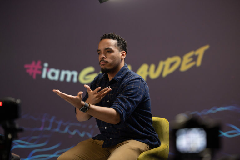 A person sitting in a yellow chair, gesturing with their hands while speaking. The background features the hashtag "#iamGalaudet" in colorful text. There are cameras set up in the foreground, indicating a recording or interview setting.
