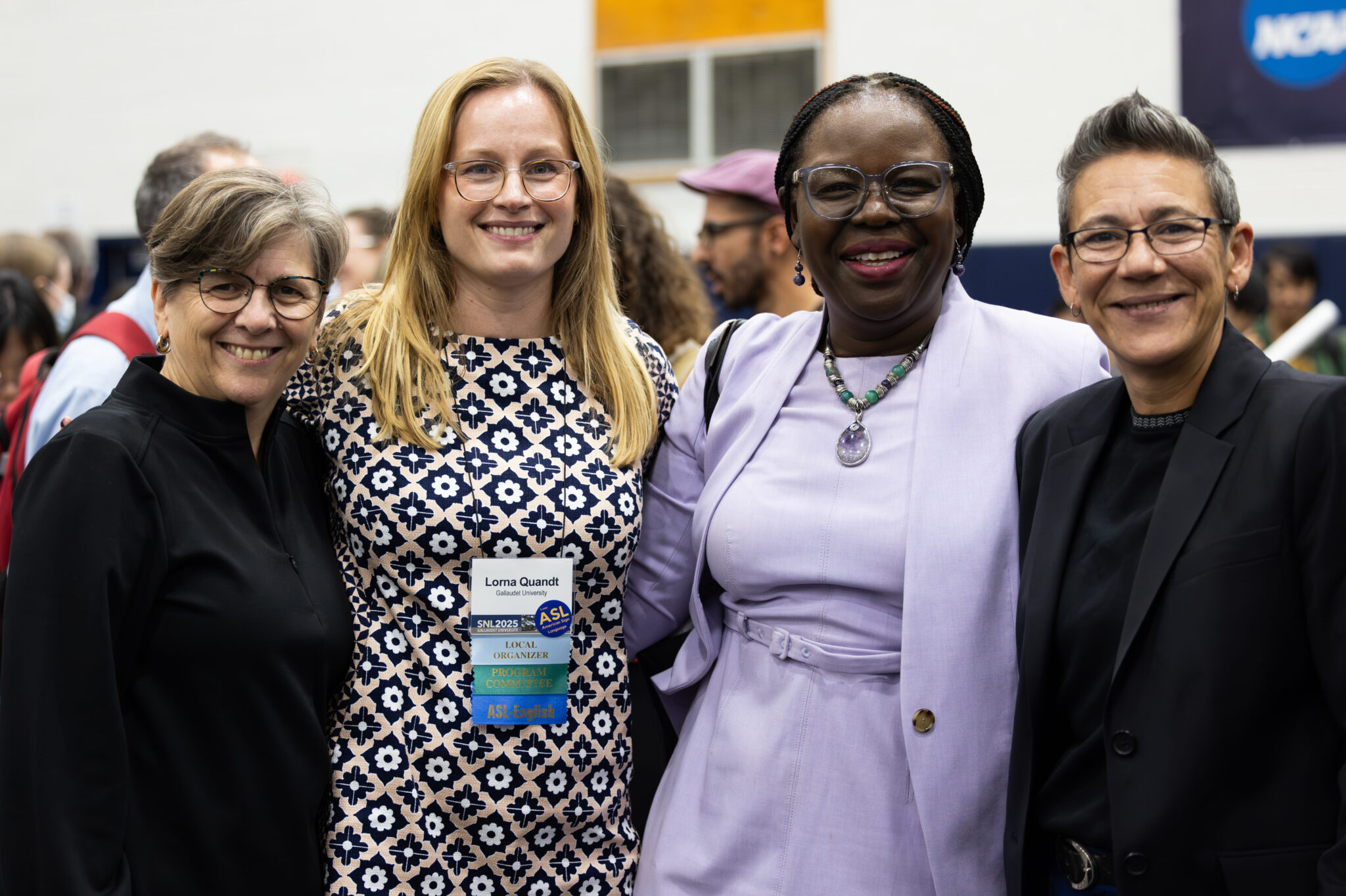 Four women pose in a row with their arms around each other. They are in a large room filled with people.