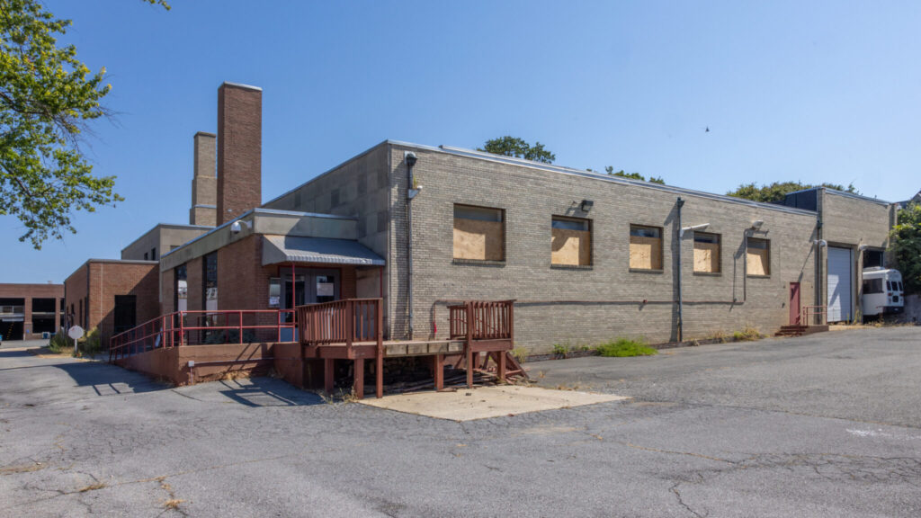 A photo of the corner of an aged, pale brick building with an industrial look. It has two chimneys in the upper left of the image. Around the corner is pavement.