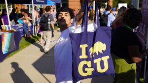 A photo of a student holding up a blue bag with yellow block graphics: "I [bison silhouette} G U". The student is partially obscured by shadow. They have tousled black hair, a mustache, and white t-shirt. In the background is a walkway with booths, tents, and mingling people.