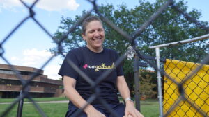 A photo of Gallaudet President Roberta Cordano smiling at the camera, through the chain link fencing of a dunk tank. She is wet from having been recently dunked in water below. She is wearing a dark blue t-shirt reading "#iamgallaudet". The Gallaudet campus is in the background, with grass, trees, and a large building.