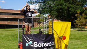 A photo of a student standing on the rim of a dunk tank after climbing up from a recent dunk. They are soaking wet. They wear a black t-shirt with a white graphic. The rim of the dunk tank has a chain link fencing, with the target below to the right in front of a yellow backdrop. The Gallaudet campus is in the background, with grass, trees, and a large building.