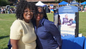 A photo of two people posing and smiling together in a light embrace. The person on the left has luxurious black curls, glasses, and a fuzzy yellow shirt. The person on the right has black hair in braids, sunglasses, and a purple shirt. Behind, a booth is visible for the Gallaudet University Department of Interpretation. They are standing outside on a sunny day. In the far background across a grassy area are tents, booths, and mingling people.