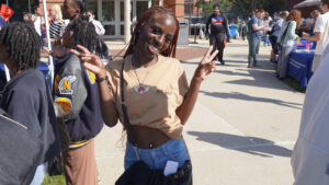 A photo of a person smiling and making peace signs to the camera. They have long braided hair, a nose stud, a necklace, and bag across their shoulders. Their tan t-shirt is pulled up in a crop top style. They are standing outside on a sunny day. In the background is a walkway with tents, booths, and mingling people.