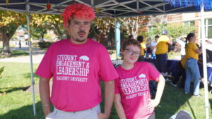 A photo of two people posing and smiling together under a tent on a sunny day. Both wear pink t-shirts that read "Student Engagement & Leadership / Gallaudet University". The person on the left is much taller, with big curly hair that is dyed orange on top. The person on the right has lightly curly short red hair and glasses. In the background are more tents, booths, and mingling people.