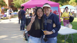 A photo of two people posing and smiling together. The person on the left is making the peace sign with their right arm. The person on the right has a Gallaudet pullover jacket and lanyard. They are standing outside on a sunny day. In the background is a walkway and grassy area with tents, booths, and mingling people.