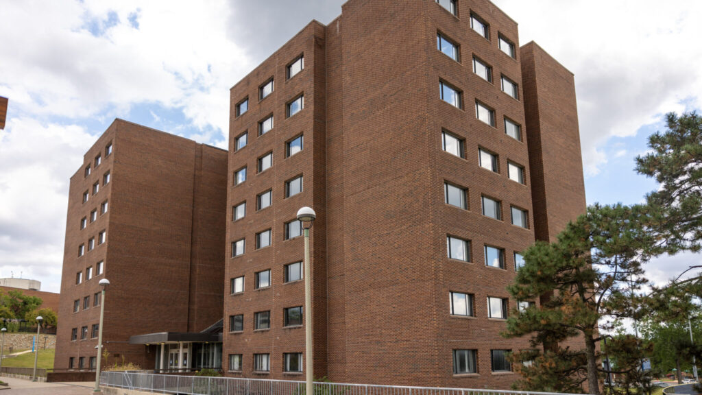 A photo looking up at a corner view of a large red brick building with two wings. One facade on each side of the corner has a grid of 18 large windows.
