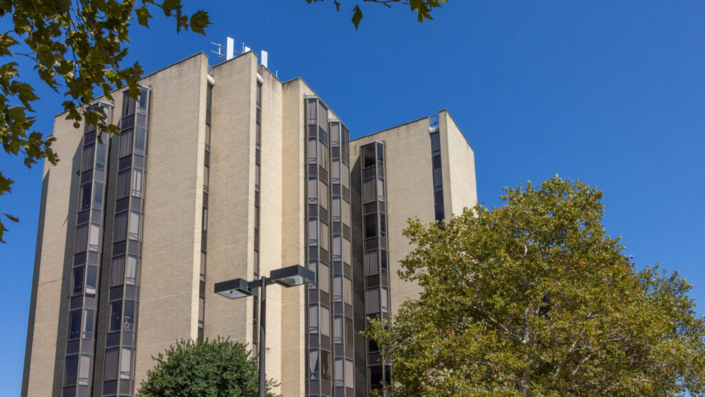 A photo looking up a tall building composed of tan stay, with columns of gray bay windows. In the foreground are trees.