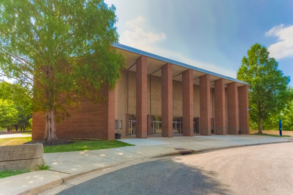 A filtered photo of a red brick building, with a facade of six columns in front of the entrance. Around the building is a grassy area with tall, bushy trees. In front is a walkway and roadway parallel to the length of the building.