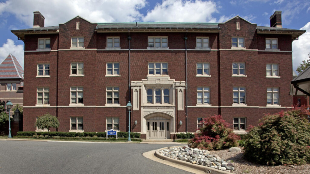 A photo of a large Victorian-style building with red brick and white windows features. The entrance is a double door surrounded by an ornate white stone feature. In front is a roadway and island with bushes.