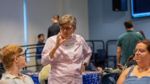 A photo of Gallaudet President Roberta Cordano standing and signing down to the left at a seated person. There is another seated person on the right. Another tables and more people are visible in the background.